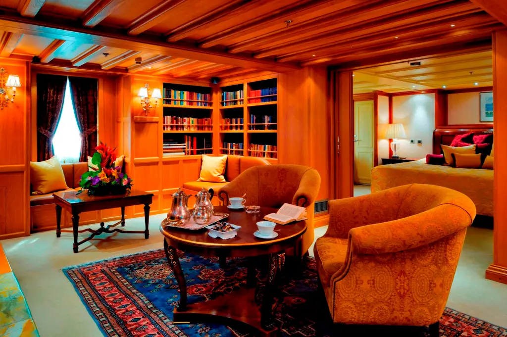 Wood-panelled sitting room with bookshelves and armchairs, a bedroom visible through the doorway