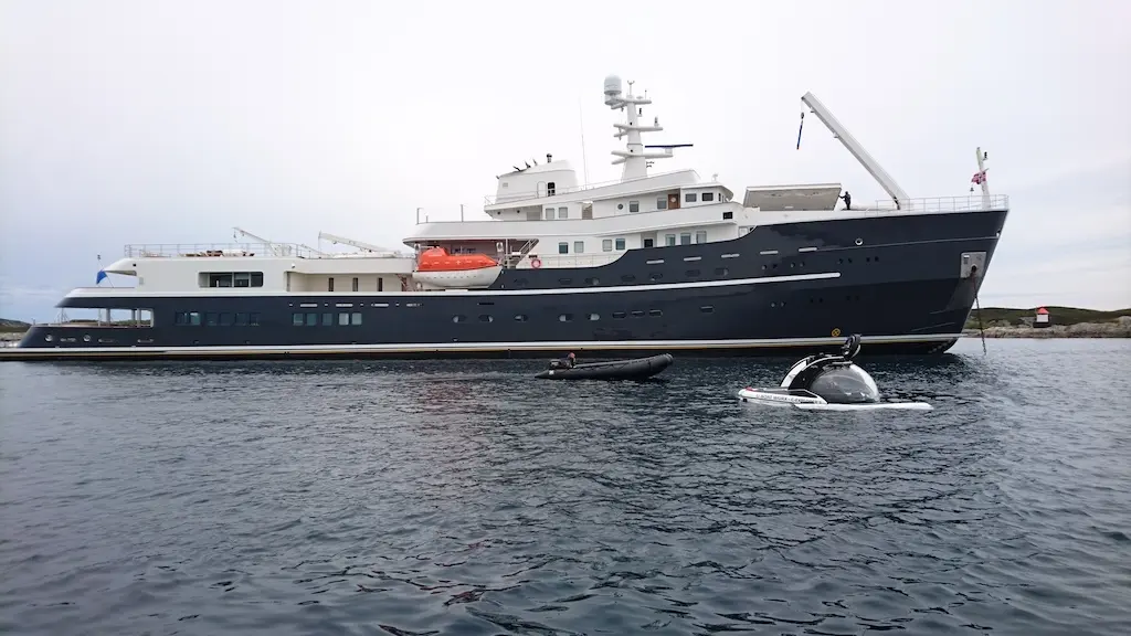 A small submarine with a clear acrylic dome surfacing beside Legend at anchor, an inflatable tender nearby and the yacht's foredeck crane raised