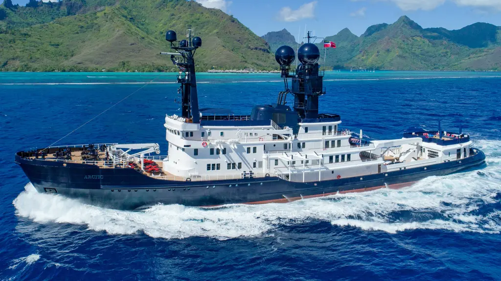 Arctic under way at speed in deep blue water, seen from the starboard side: black hull, white stepped superstructure, two black masts carrying radar domes, and steep green peaks behind