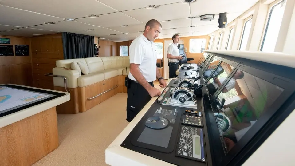 Two officers at the bridge console of Arctic, with engine controls, screens and a chart table, and a sofa at the back of the wheelhouse
