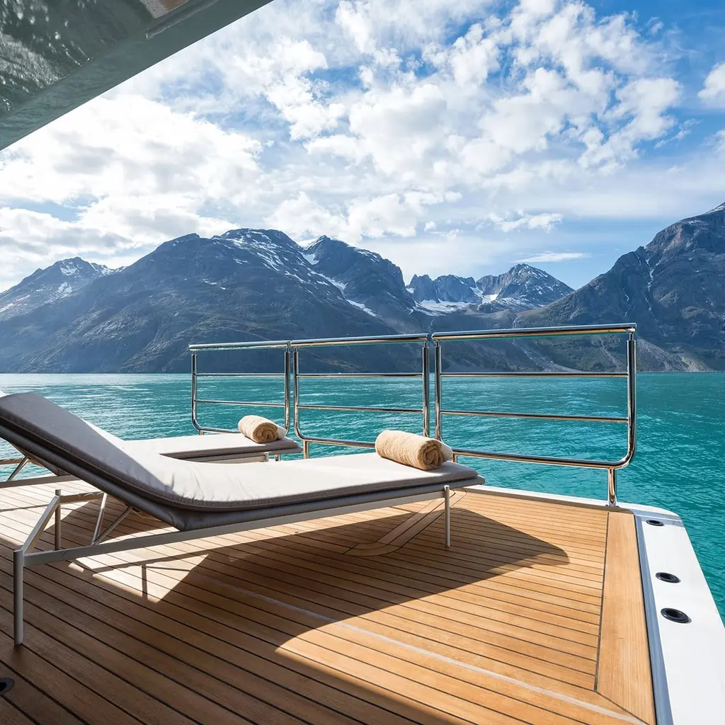 A sun lounger on a teak deck behind a rail, facing a fjord and snow-capped mountains