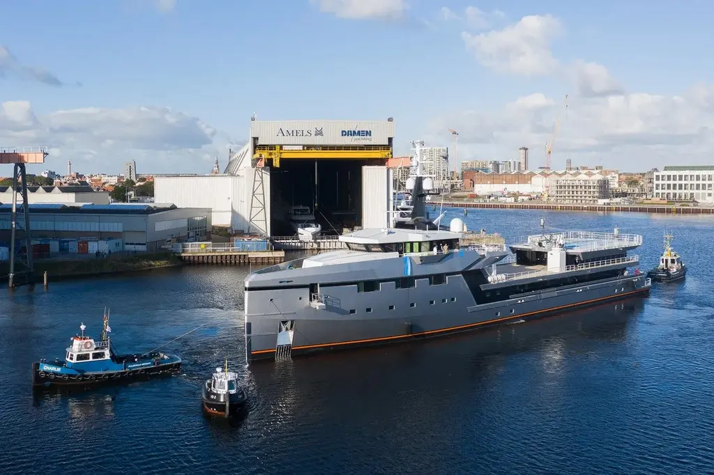 Abeona being moved by three tugs in a shipyard basin in front of a large shed marked Amels and Damen