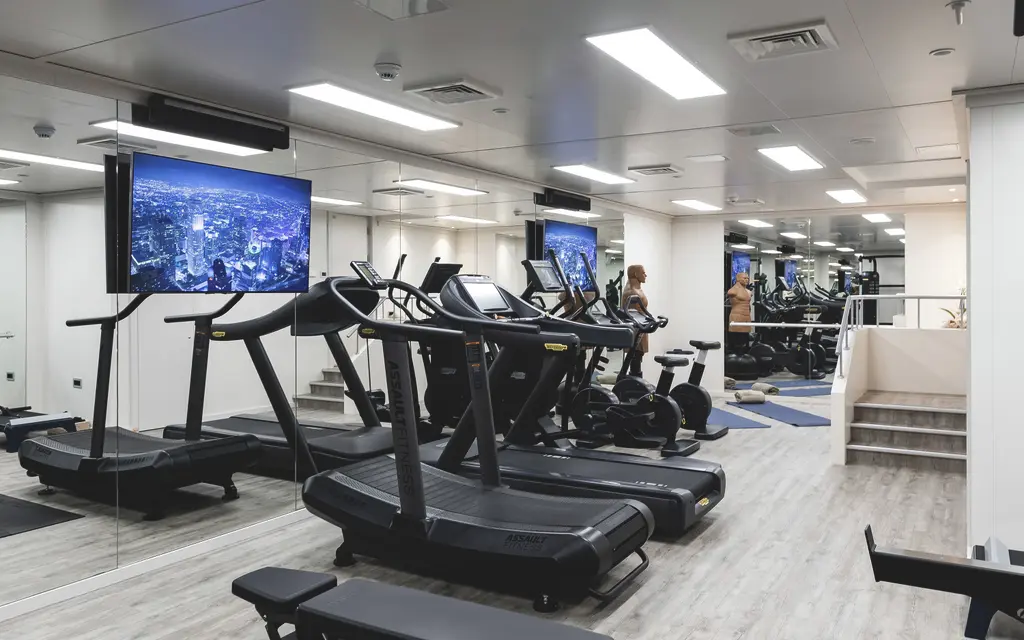 A gym with treadmills and exercise bikes facing a mirrored wall and screens