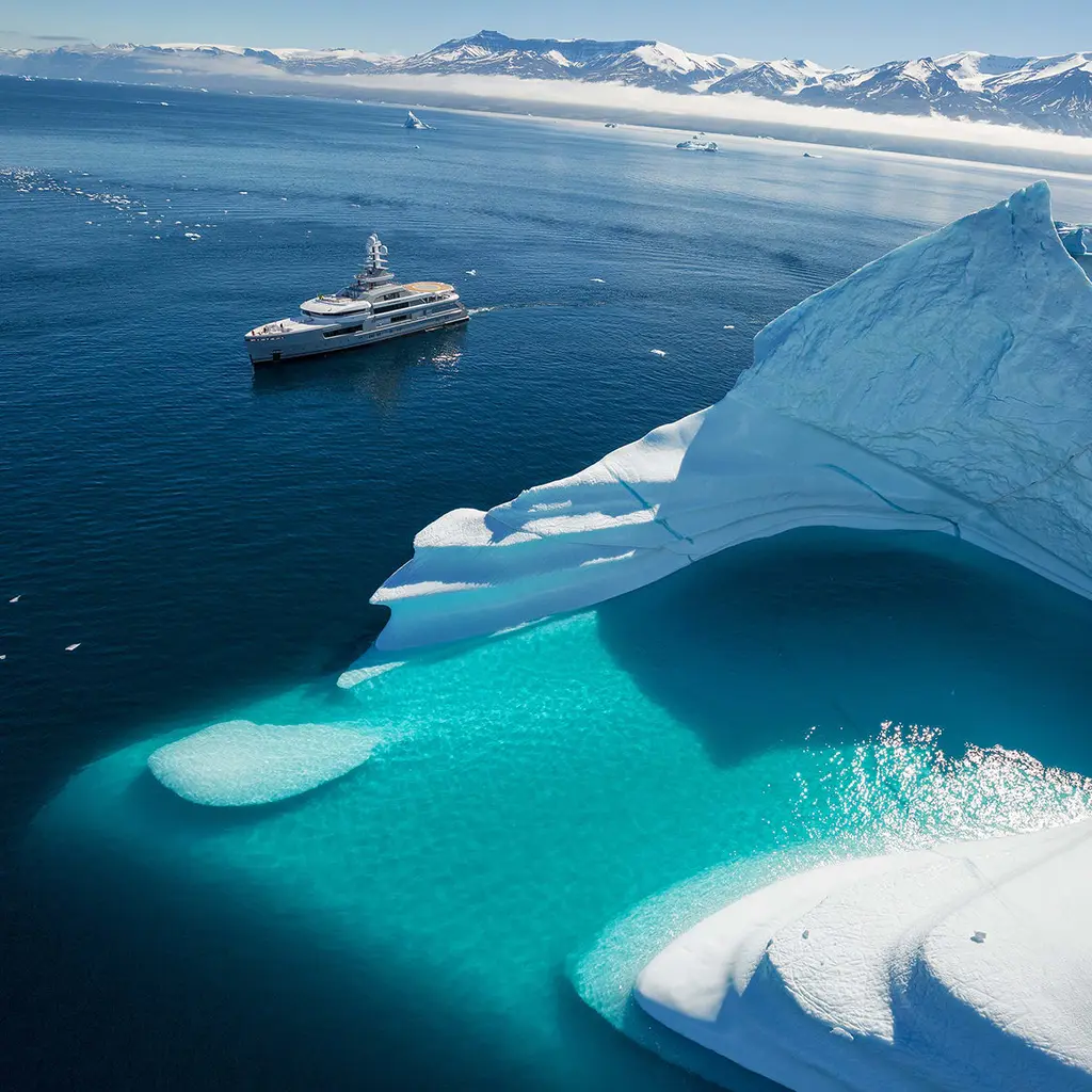 High aerial view of a large turquoise iceberg in the foreground, with Cloudbreak small in the distance against a mountain coast