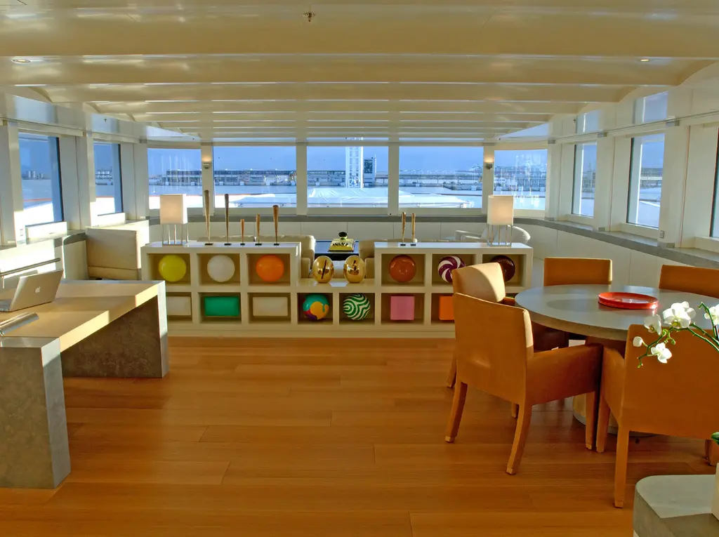 A bright lounge with windows on three sides, an oval table with orange chairs and a coloured display cabinet