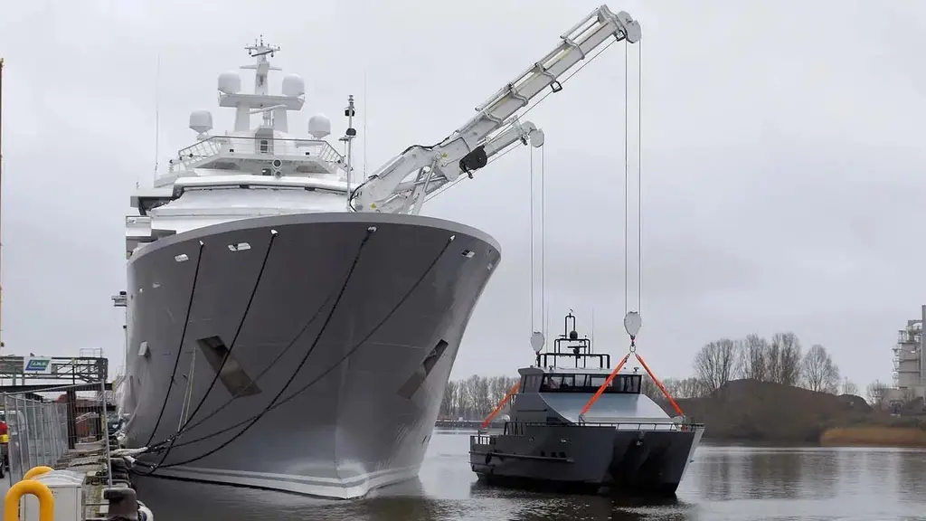 Andromeda's bow alongside a quay, with the foredeck cranes lifting a dark grey tender from the water
