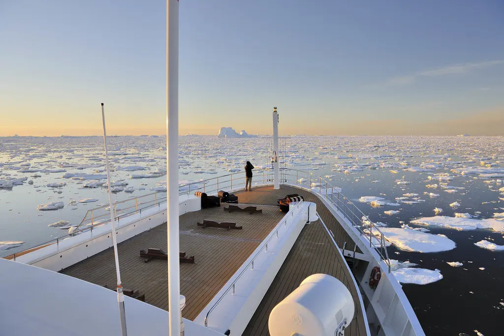 View forward from the bridge deck over Legend's teak foredeck, with a person at the bow rail and sea ice and an iceberg on the horizon under a pale evening sky