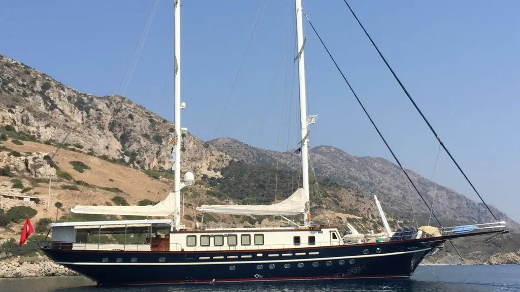 Sea Dream at anchor below a rocky hillside, sails furled, side profile