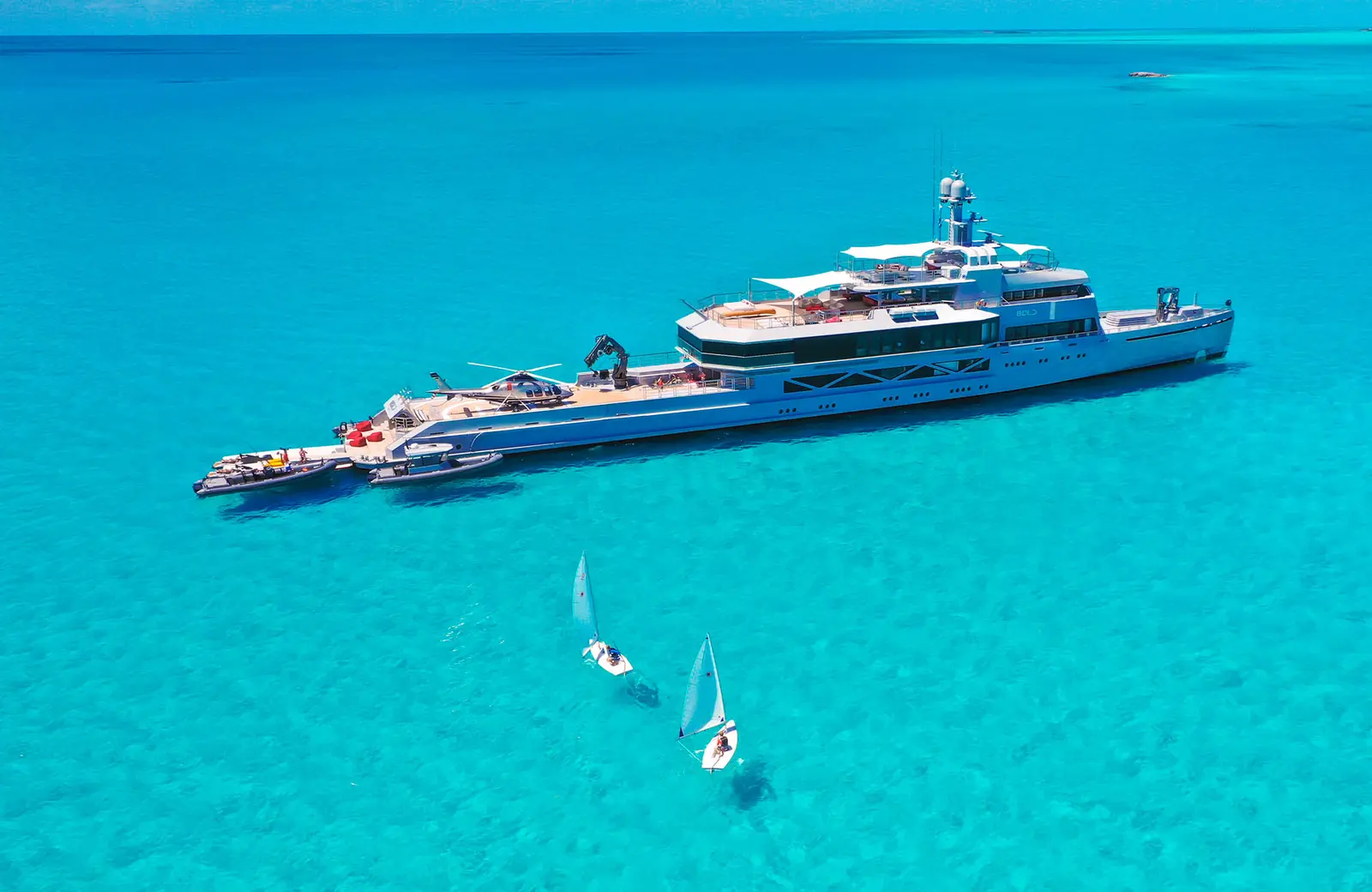 Aerial view of Bold at anchor over pale turquoise shallows, with two small sailing dinghies in the foreground