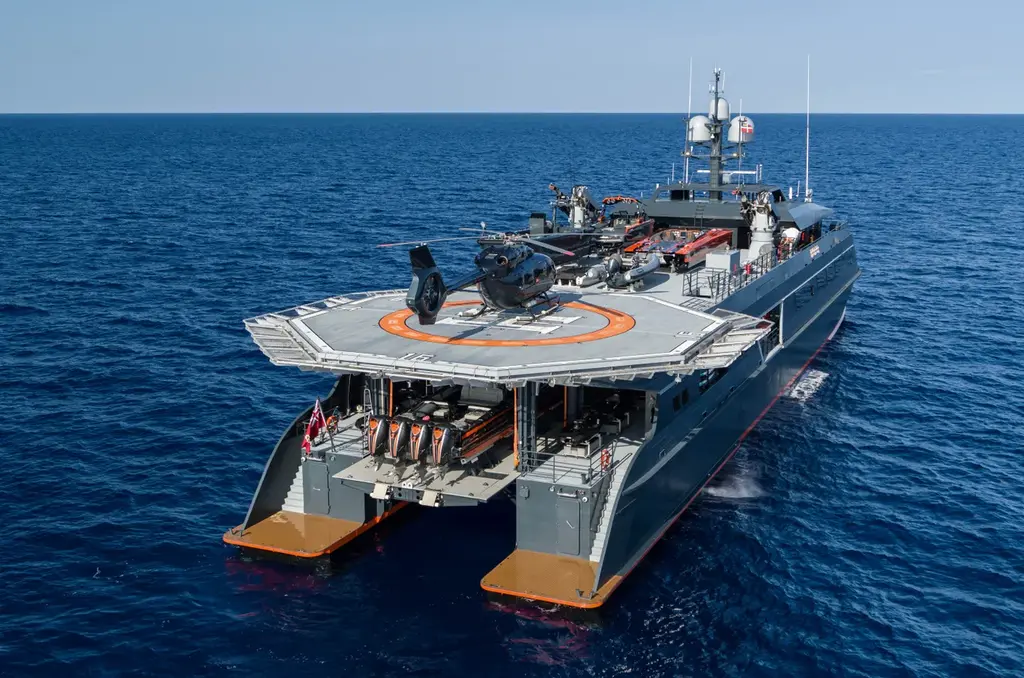 The stern of Hodor at sea: the helipad with her helicopter overhangs the open space between the two hulls, with a centre-console boat with four outboard engines stowed underneath on the main deck