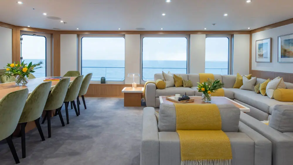 A saloon with large windows onto the sea, a long dining table with green upholstered chairs on the left and a pale grey L-shaped sofa with yellow cushions on the right