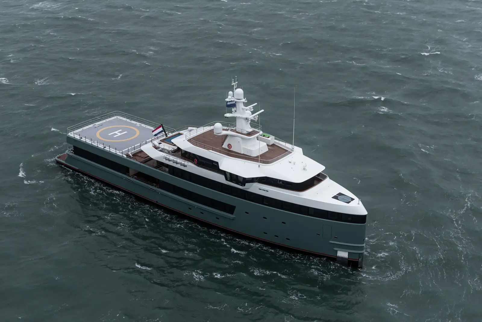 Aerial view of Anawa under way on grey-green water, seen from above her starboard side: a green-grey hull, white superstructure and a helideck with an orange landing circle over the stern, a Dutch flag on the aft mast
