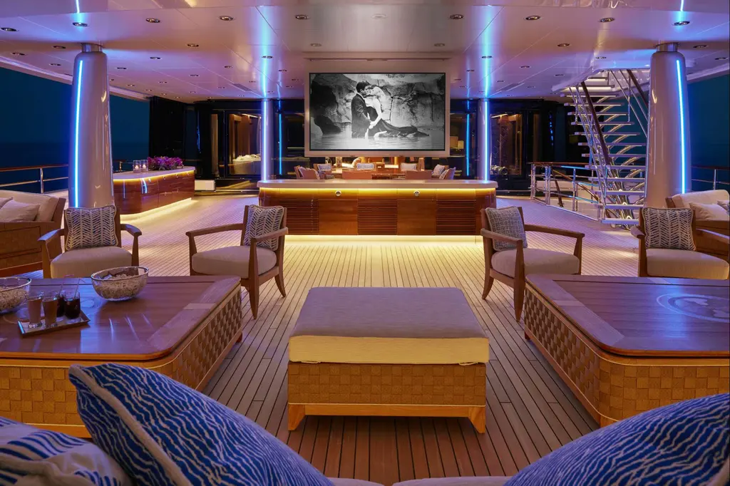 Covered teak deck at dusk with armchairs, low tables and a large film screen, lit in blue