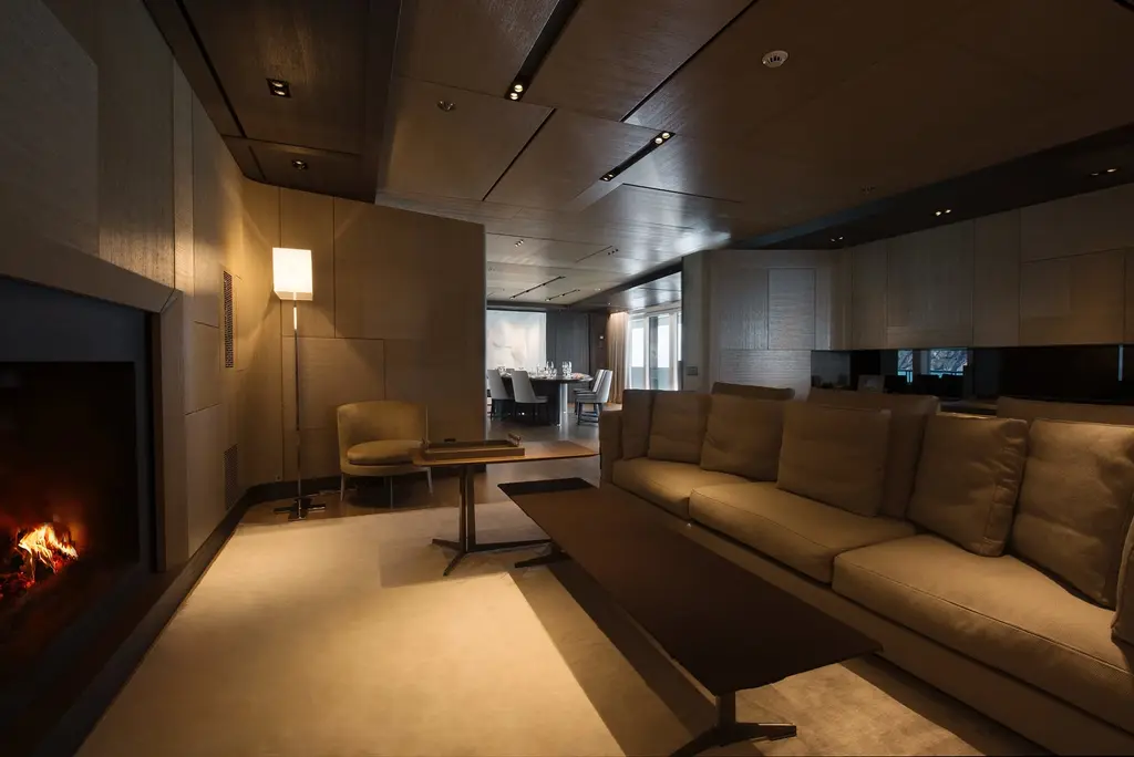 A saloon with a lit fireplace set in a dark wall, low brown sofas and a reading lamp