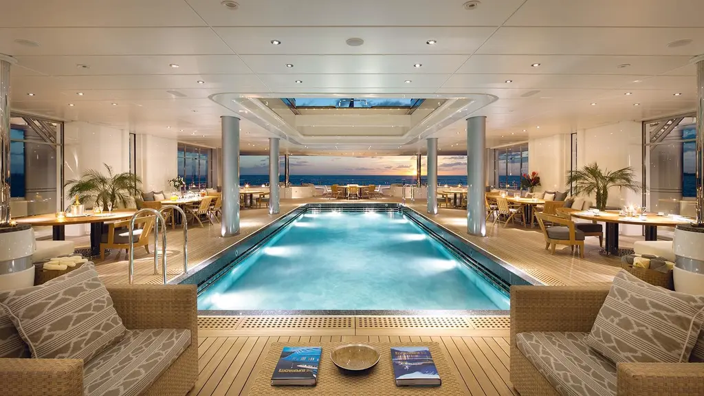 Covered pool deck with a long lit pool, tables on both sides and a glass roof panel overhead