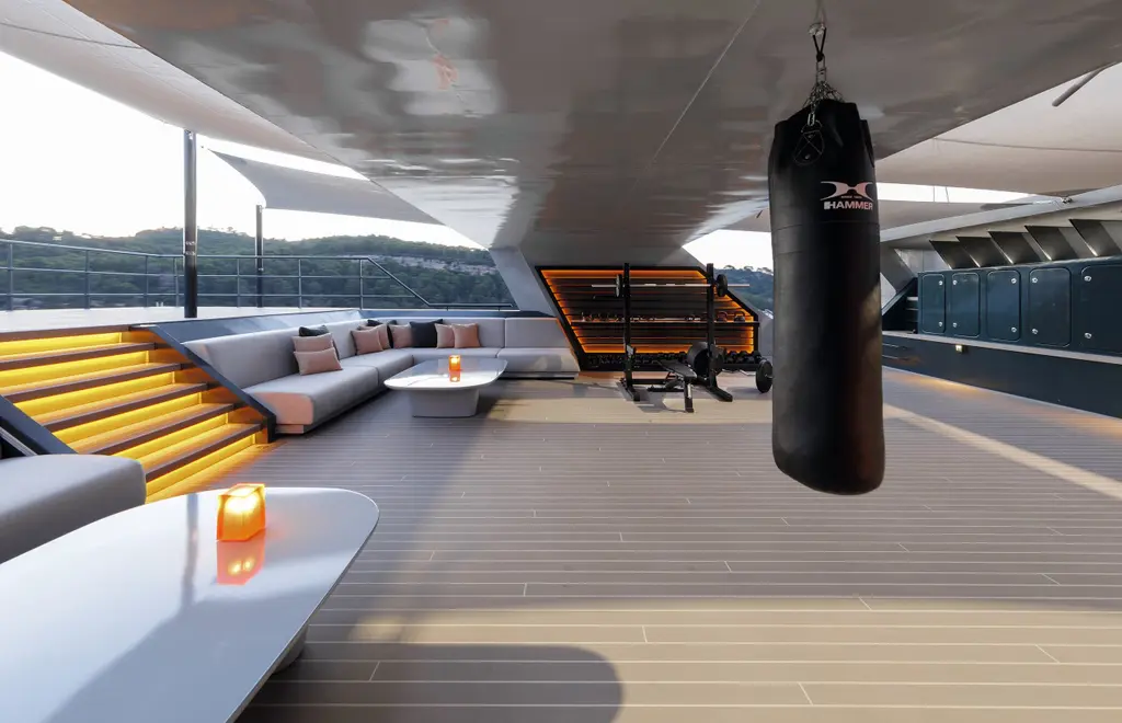 Nebula's sundeck under a white arch: a grey L-shaped sofa, a punch bag and fitness equipment, with a coastline beyond the rail