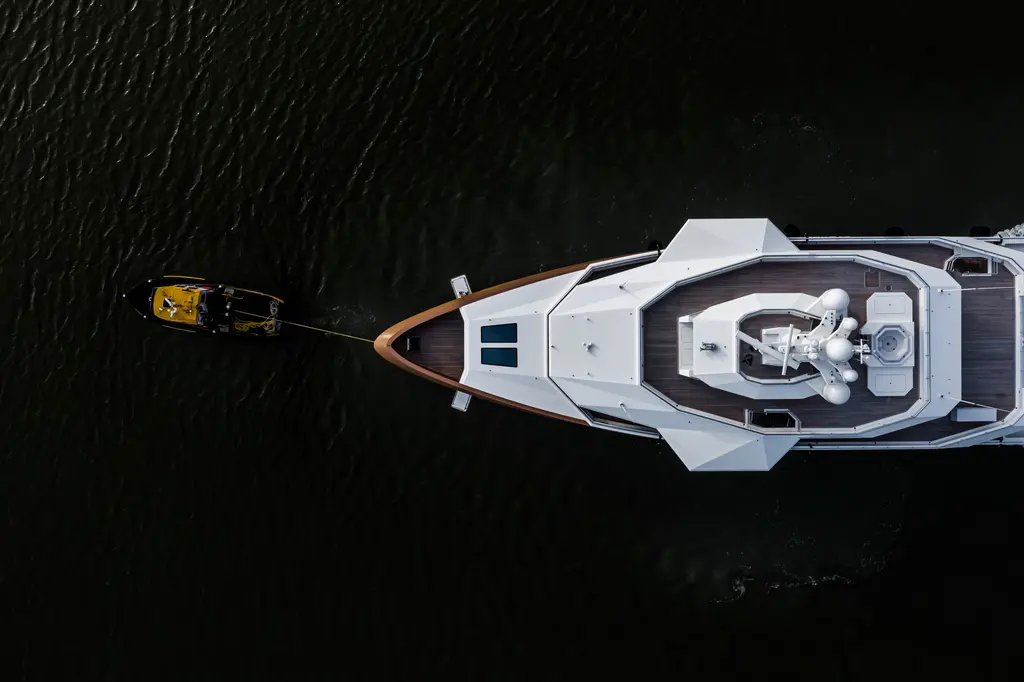View straight down on La Datcha's bow and foredeck over dark water, with a small yellow boat on a line ahead
