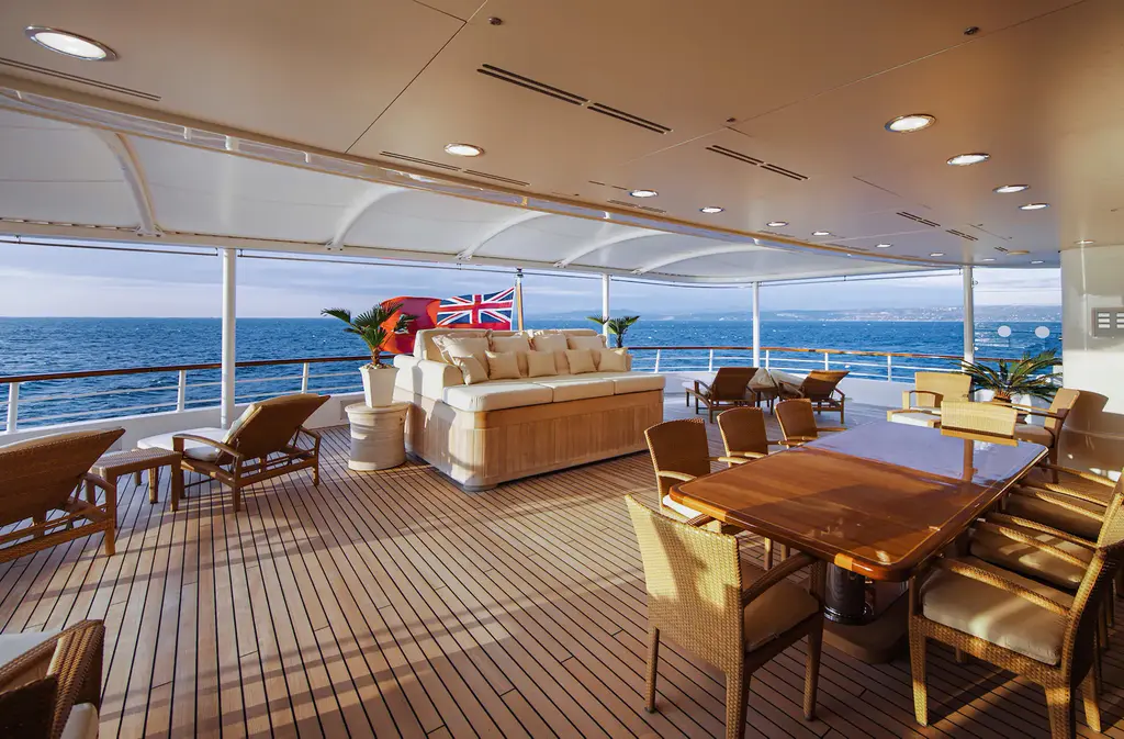 Covered aft deck with teak floor, wicker sofa, dining table and loungers, sea behind