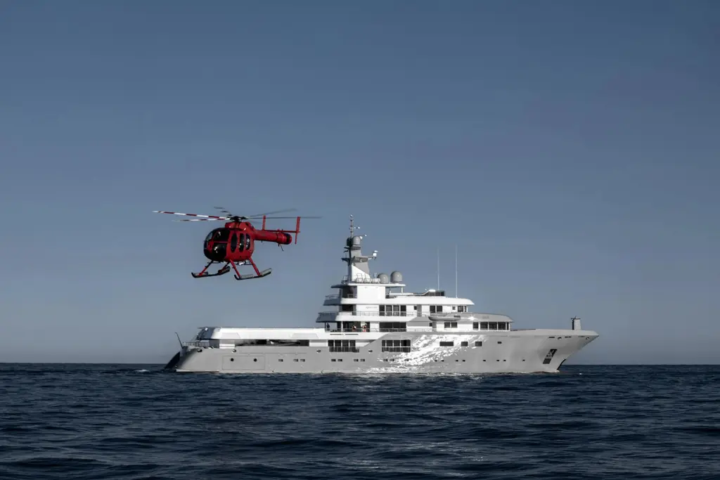 A red helicopter flying alongside Planet Nine at sea, with the yacht in profile below it