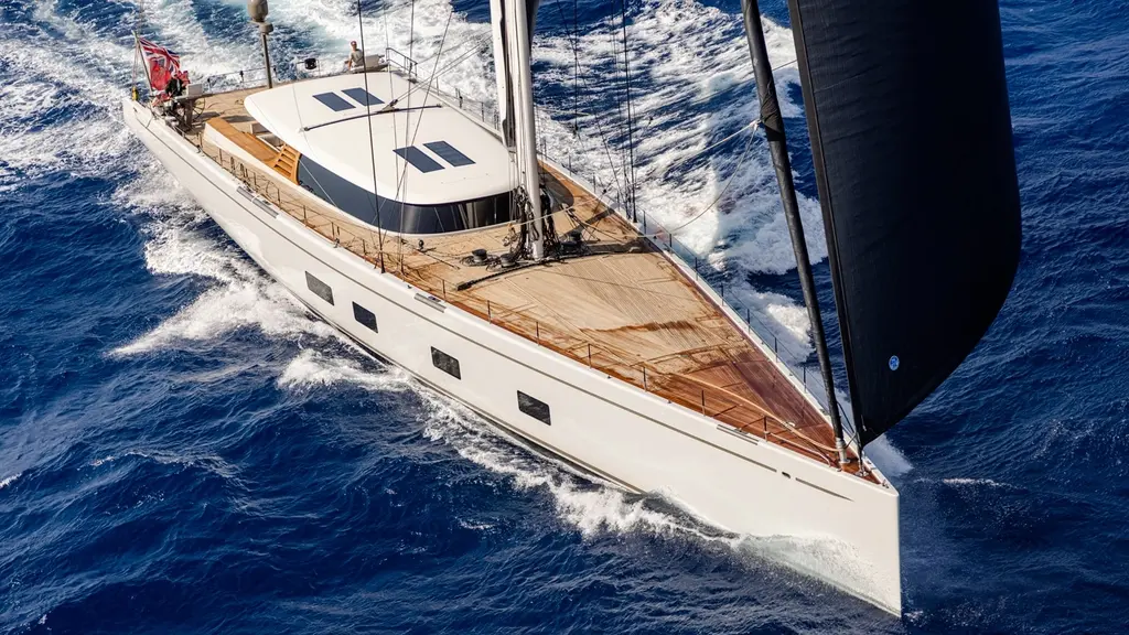 Aerial three-quarter view of Canova sailing fast on dark blue water, white carbon hull, teak deck and dark sails