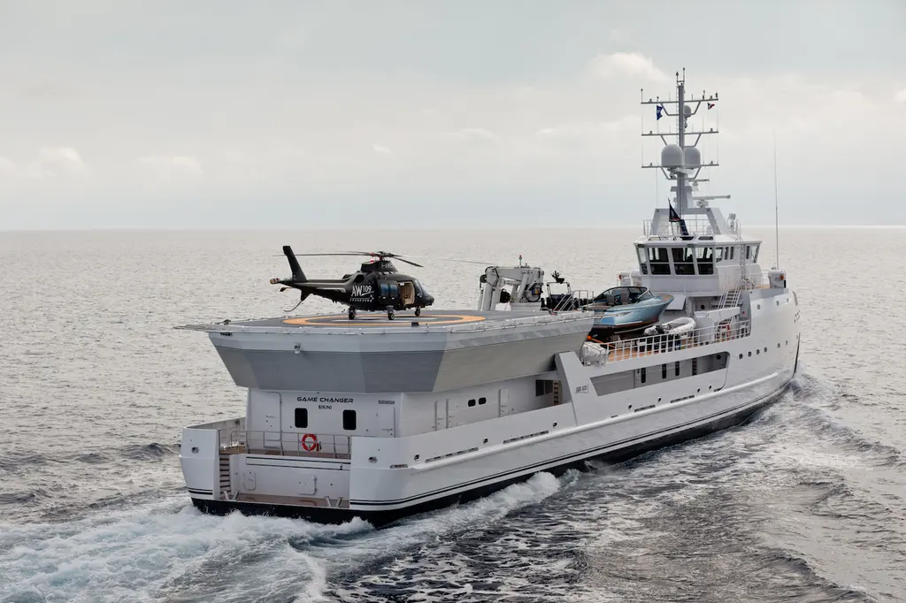 Game Changer at sea seen from the starboard quarter, with a helicopter parked on the raised helideck above the stern and a blue-grey tender on the working deck behind it