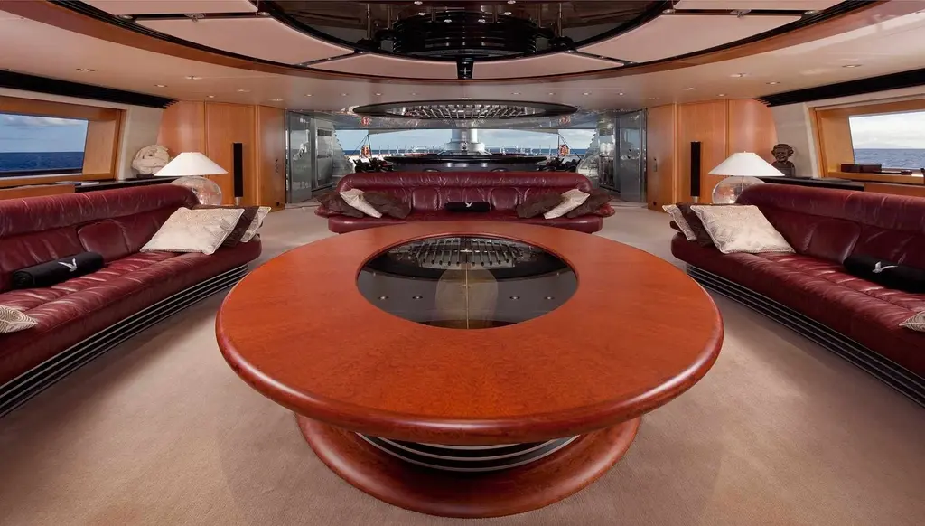 Main saloon with a round table, red sofas along both sides and windows at the far end