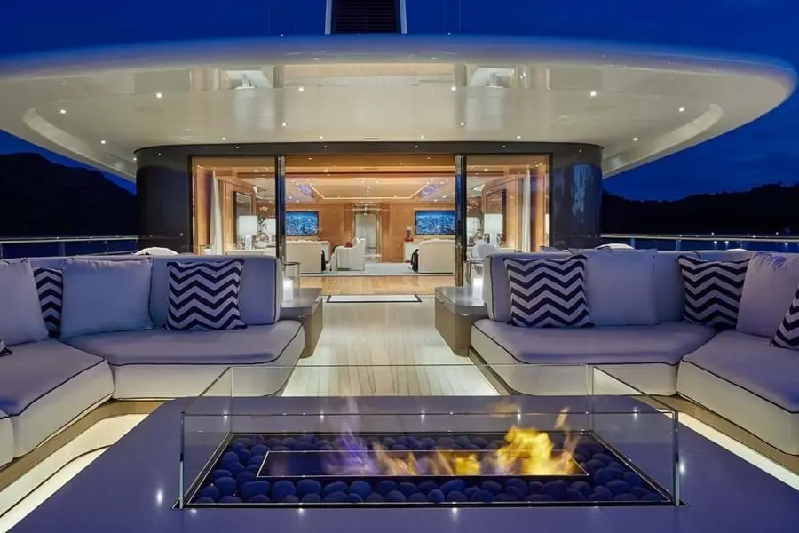 Outdoor lounge with sofas around a glass-sided fire pit at night, the lit saloon visible behind