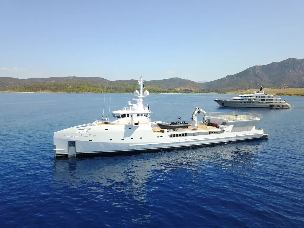 Game Changer at anchor in a calm bay with hills behind, a tender and crane on her working deck and a larger white yacht further back