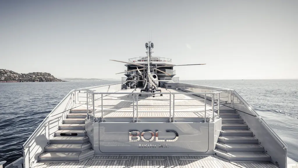 The stern from the water, with the name BOLD on the transom, stairs on both sides to the aft deck and a helicopter parked above