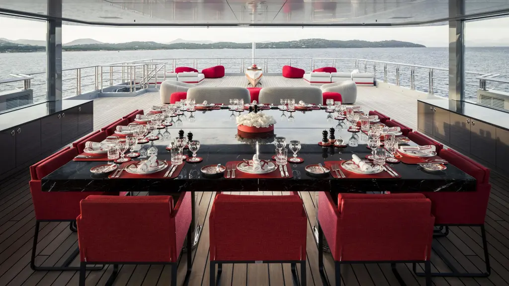 A long table laid for dinner with red chairs in the winter garden, with glass walls looking out to sea
