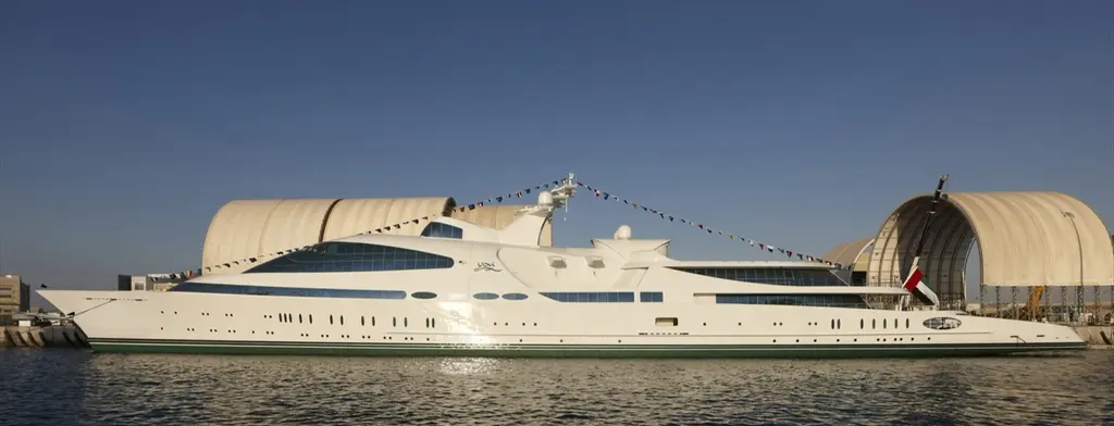 Yas in profile alongside a quay, dressed with signal flags and the UAE flag, with large arched sheds behind her