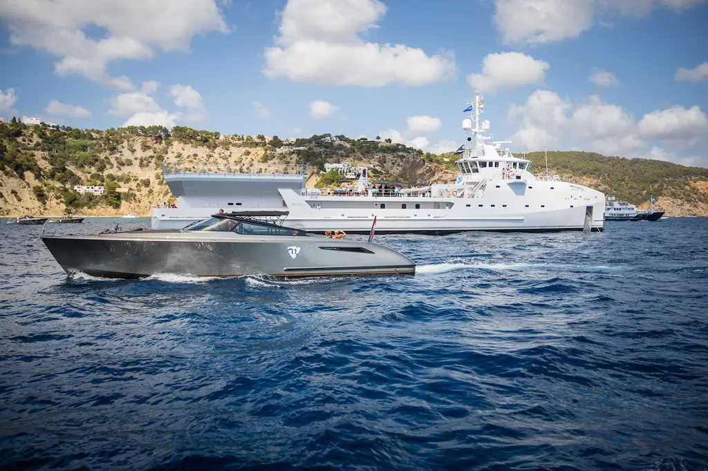 A dark grey dayboat tender running in front of Game Changer, which lies at anchor off a rocky wooded coast