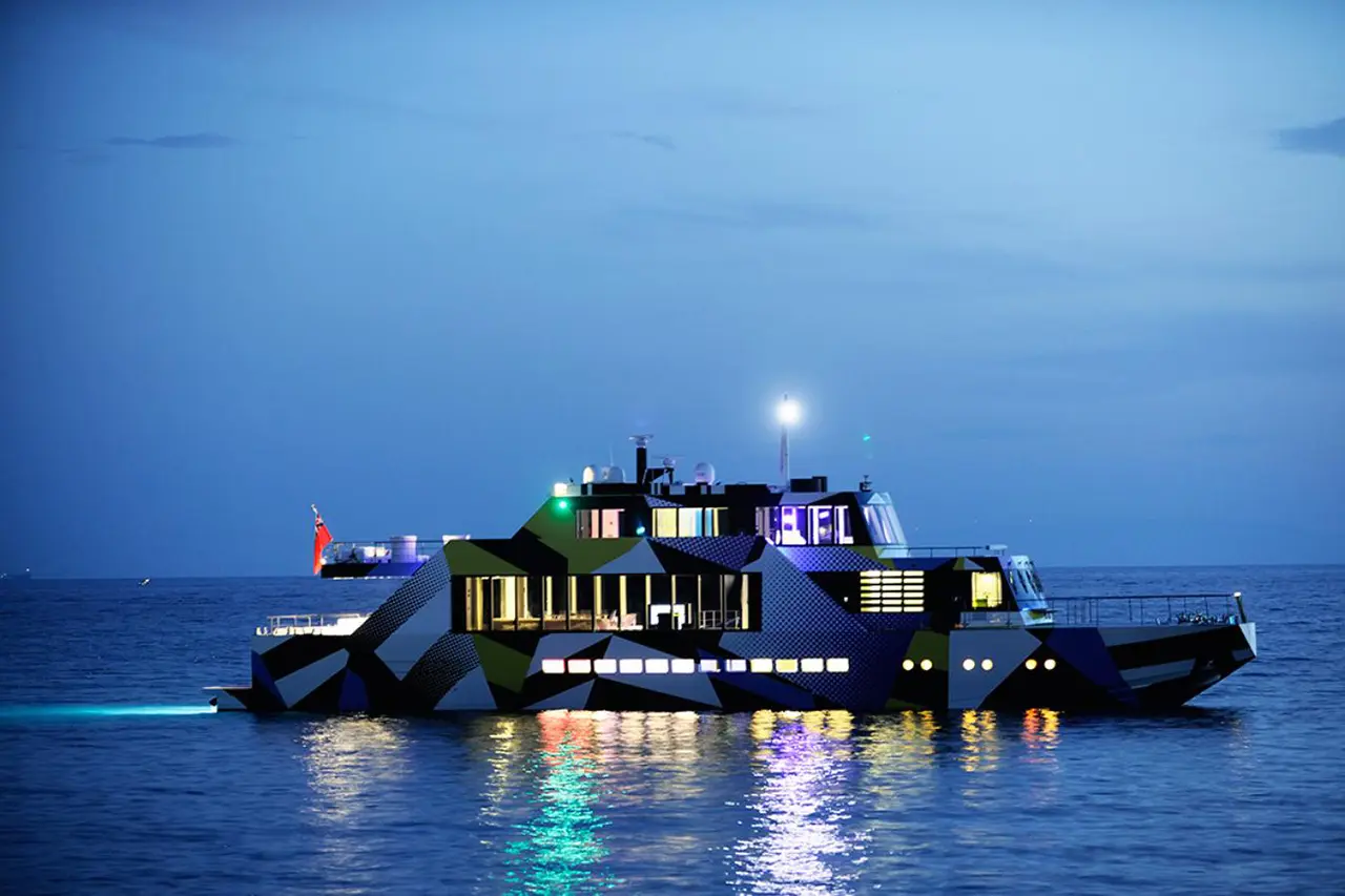 Side profile of Guilty at dusk on calm water, lit windows across two decks and the camouflage pattern on the hull