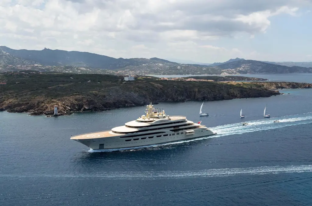 Dilbar underway past a rocky headland and a small lighthouse, with sailing yachts behind