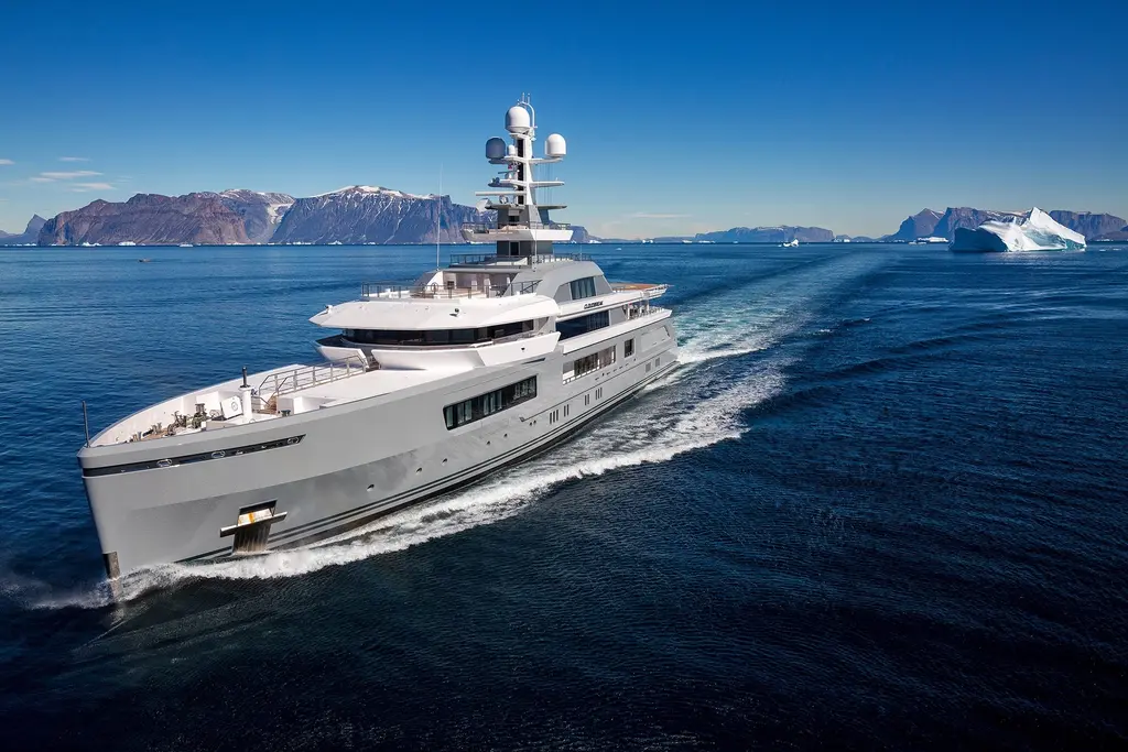 Cloudbreak under way at speed in deep blue water with a white wake, a rocky coastline and small icebergs behind