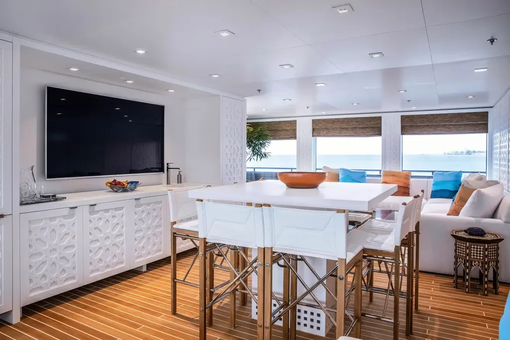 A white lounge and dining area with a large wall screen, a long high table with white chairs and sofas by wide windows