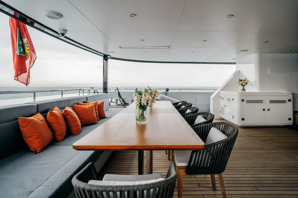 Covered aft deck with a long table, a banquette with orange cushions and a view over the sea