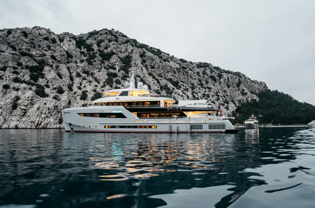 Heeus in profile at anchor in a still bay below a rocky hillside
