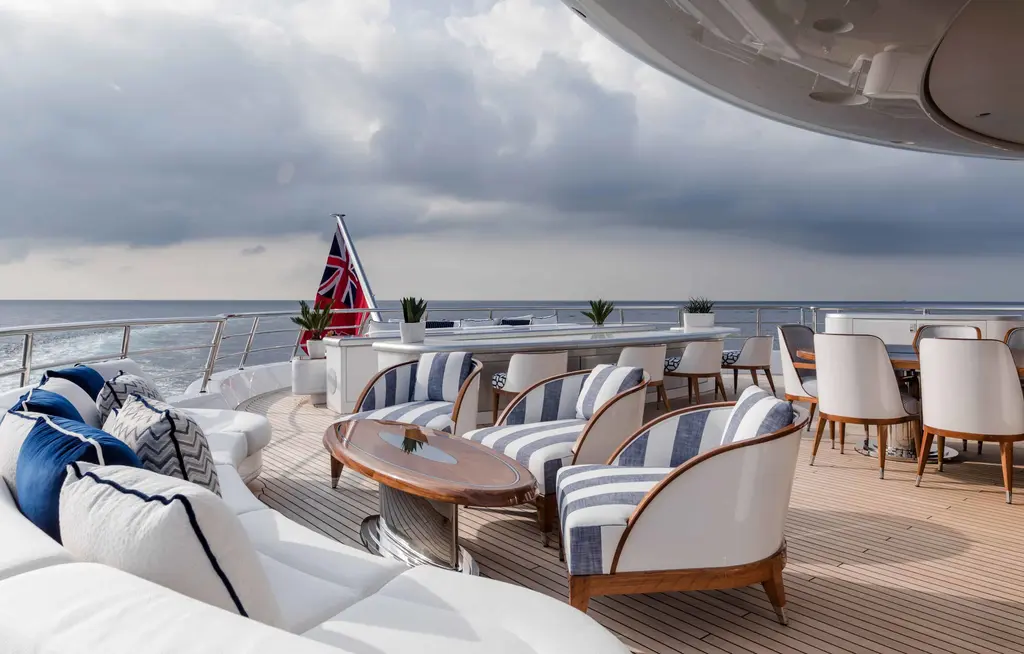 Aft deck with striped armchairs, a white sofa with blue cushions and a dining table, the sea beyond the rail