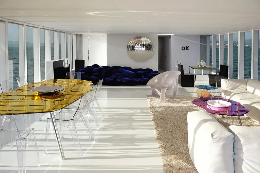 White saloon with floor-to-ceiling windows, a gold-topped dining table with clear chairs, a deep blue sofa and a mirrored wall piece