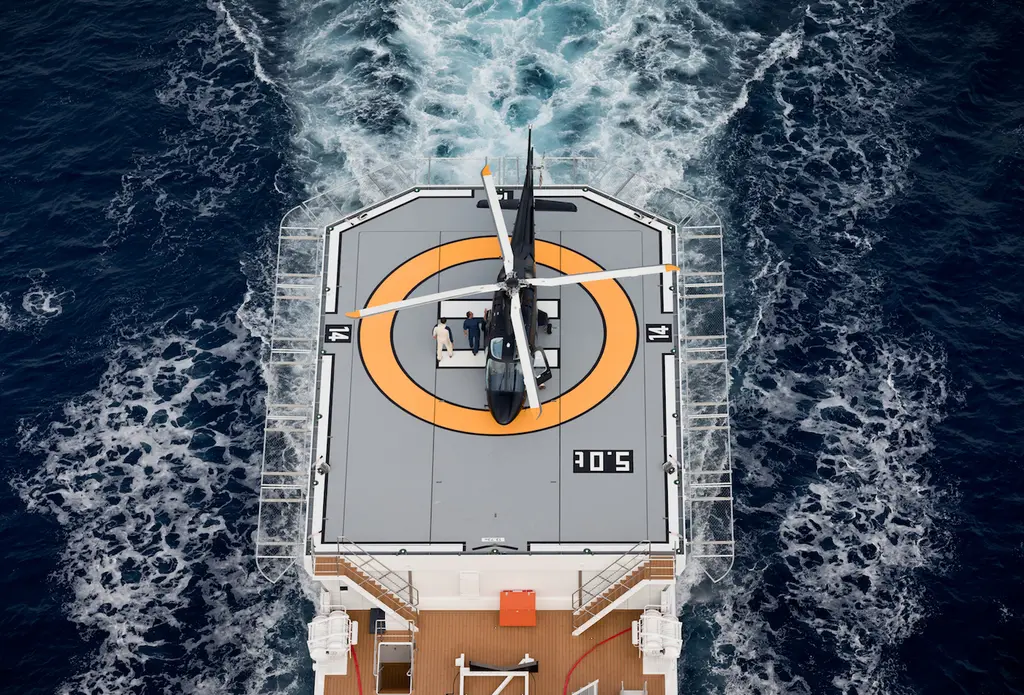 Looking down on the aft helideck of Game Changer at sea, with a helicopter parked on the orange touchdown circle and the wake spreading behind