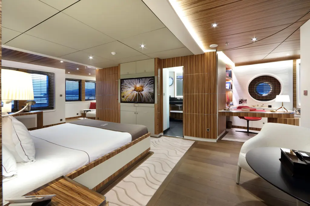 Stateroom with a double bed, a wall screen, a desk under a round porthole and an en-suite door