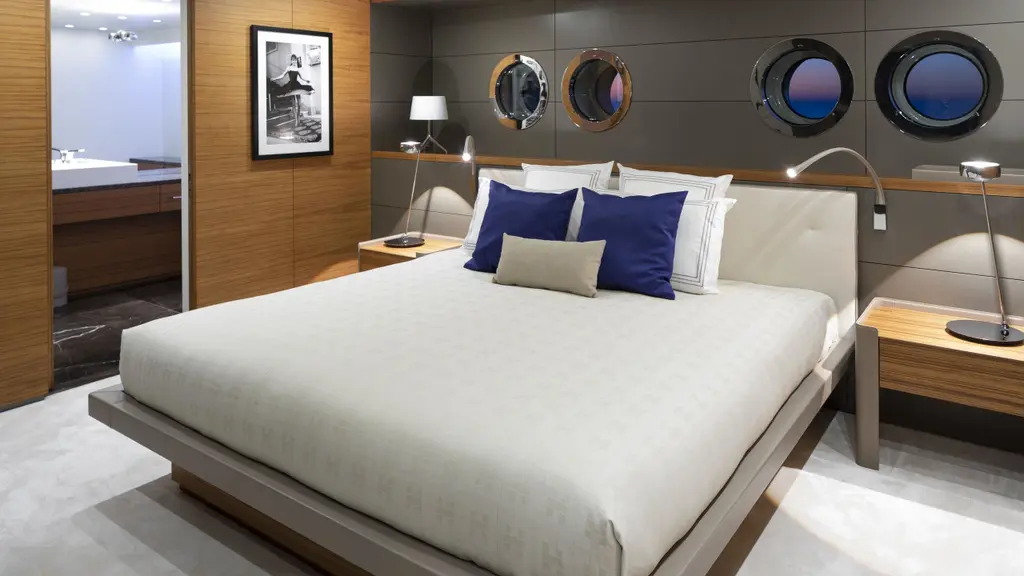 A cabin with a double bed under round portholes framed in stainless steel, with blue cushions and dark panelling