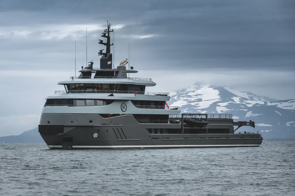 Ragnar at sea seen from the side, a grey hull and superstructure with an R emblem, in front of a snow-capped mountain under a low sky