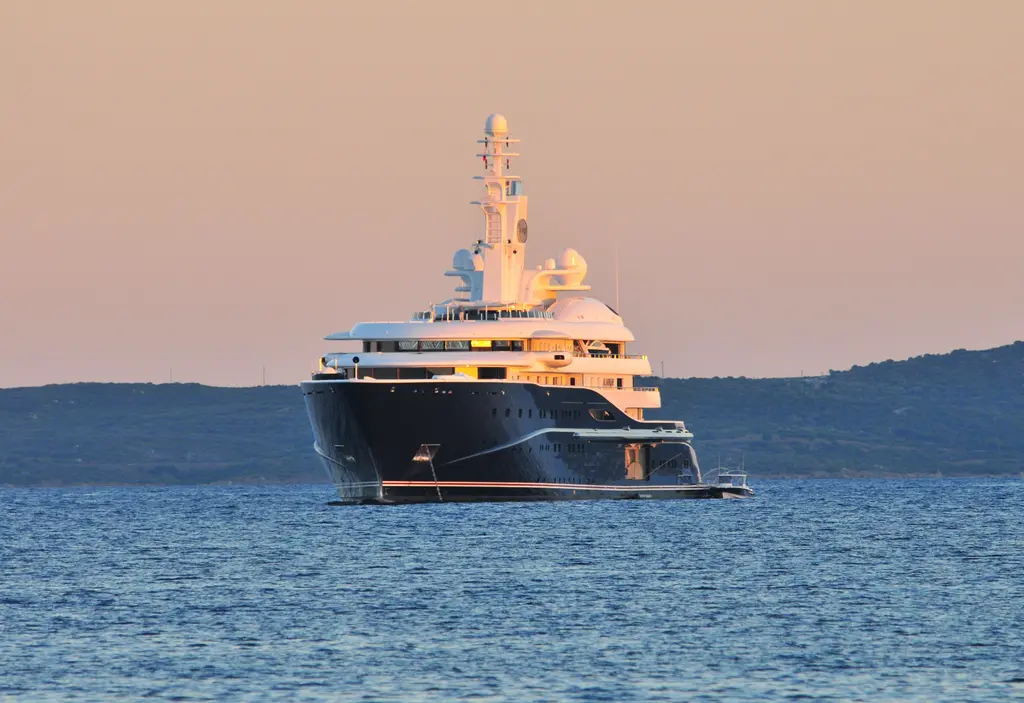 Al Mirqab bow-on at anchor in evening light, with a hilly coast behind
