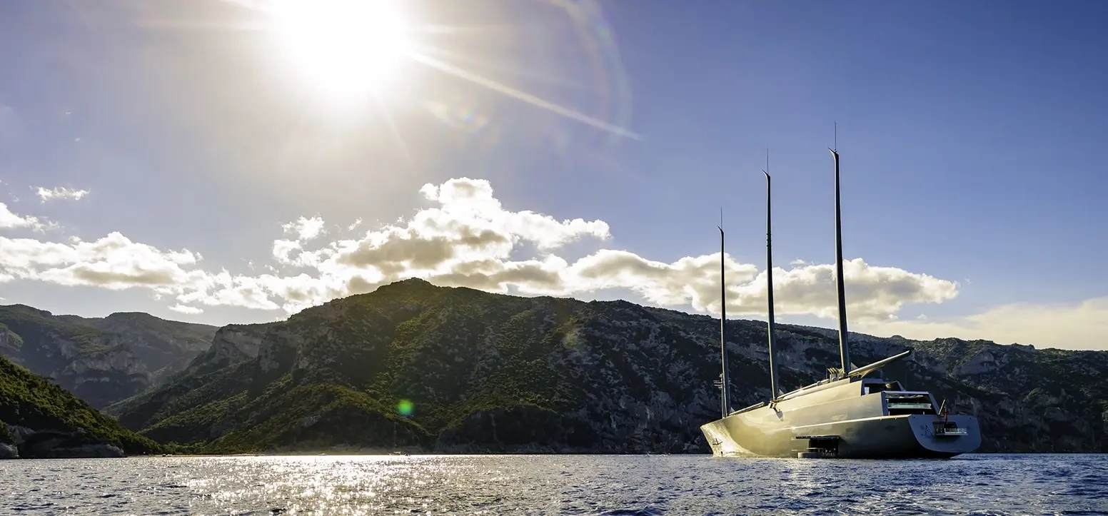 Sailing Yacht A at anchor below green mountains, sun low over the bay