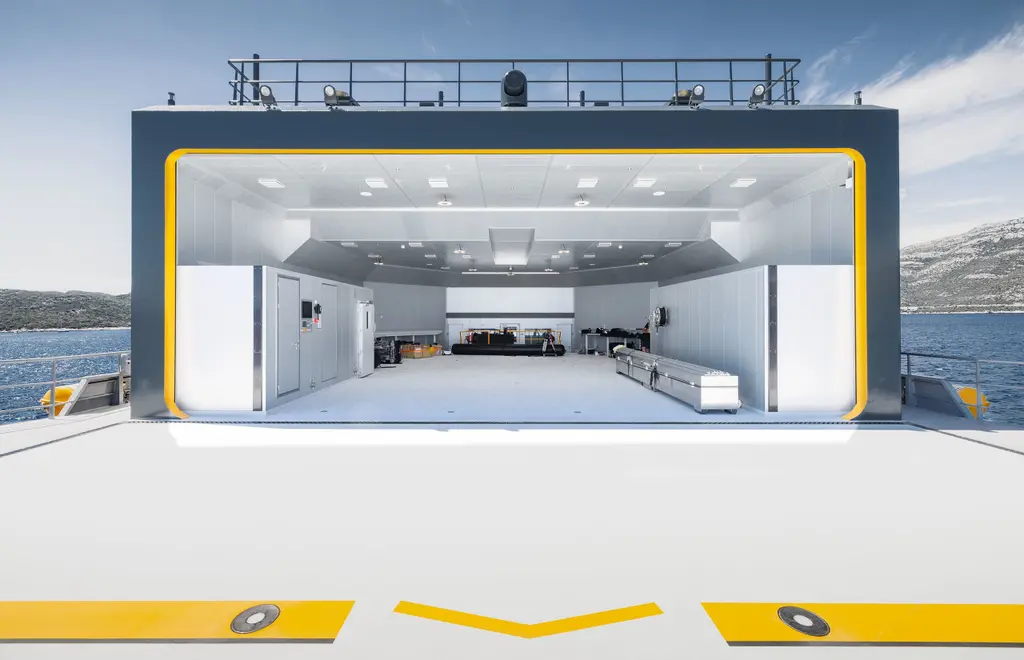 Looking from the helideck into Nebula's empty helicopter hangar, a white box framed in orange with ceiling lights and workbenches, and the orange deck markings in the foreground