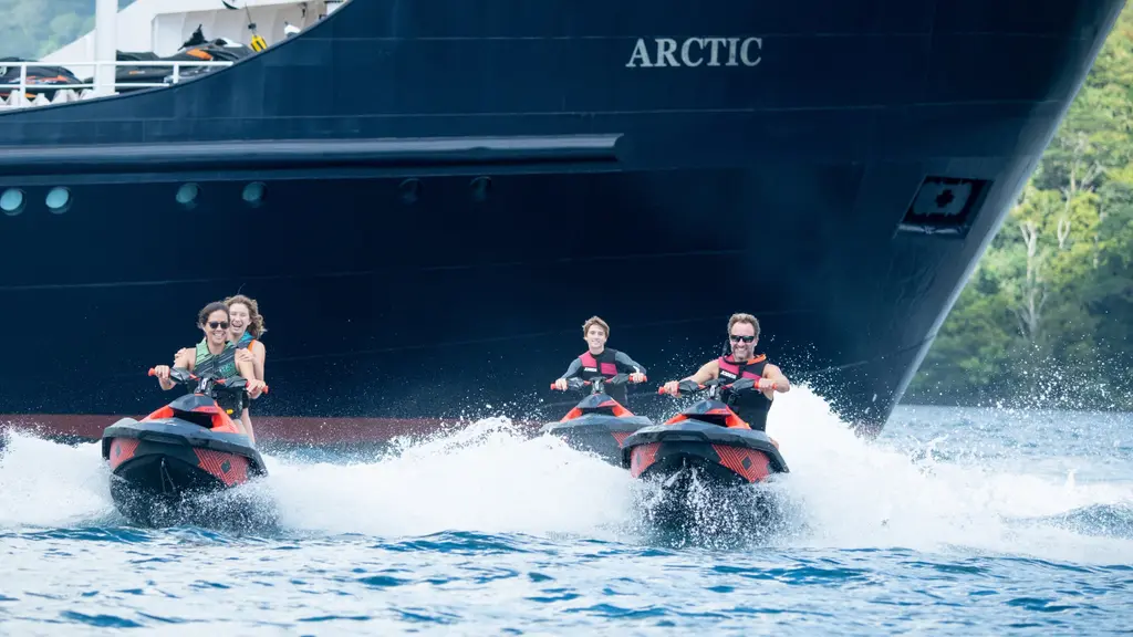 Three jet skis speeding past the black bow of Arctic, her name in white letters on the hull
