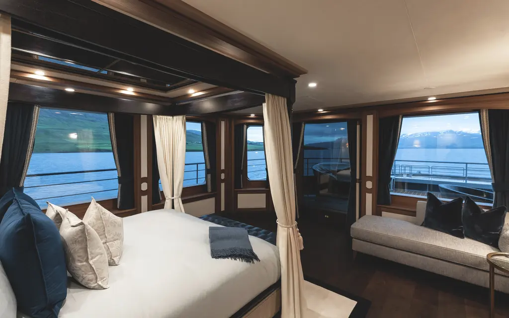 A cabin with a double bed under curtained wooden posts and windows on two sides looking out to sea and mountains