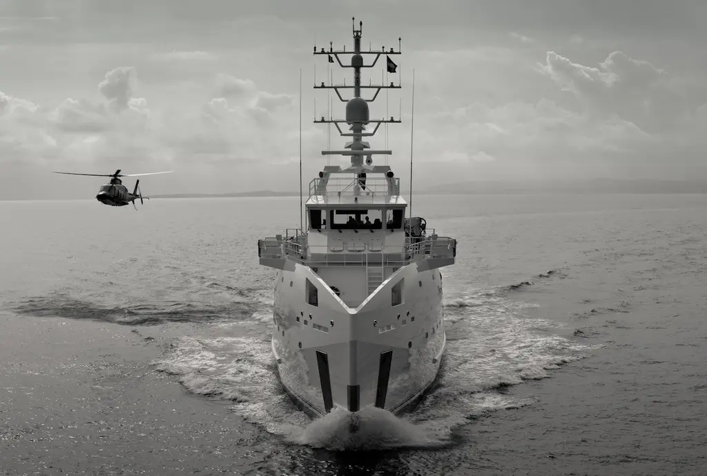 Black and white photo of Game Changer seen head-on, showing the sharp vertical axe bow, the tall mast and a helicopter flying to the left